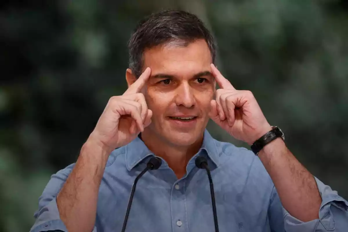 pla mig de Pedro Sánchez parlant davant de dos micròfons amb cara somrient i amb els dos dits índex recolzats al front
