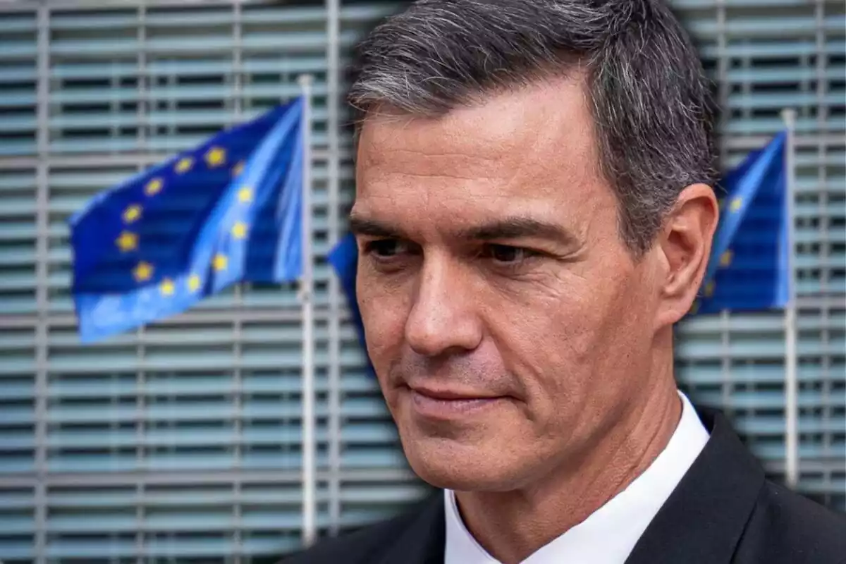 Primer pla de Pedro Sánchez amb cara seria mirant cap a terra i de fons una imatge de la façana de la Comissió Europea i diverses banderes de la Unió Europea