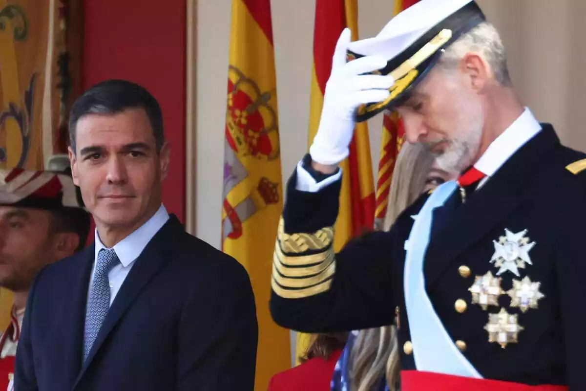 Pla mitjà de Pedro Sánchez amb un lleuger somriure mentre el rei Felip vestit de militar es col·loca bé la gorra mentre mira a terra