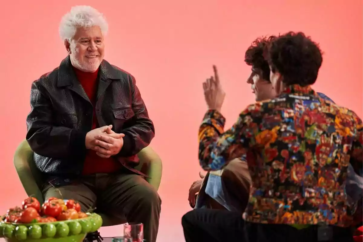Pedro Almodóvar de cabells canosos i jaqueta negra somriu mentre conversa amb Javi Calvo i Javi Ambrossi davant seu, una d'elles porta una jaqueta colorida i hi ha una fruitera amb tomàquets sobre la taula, tot en un ambient amb fons rosa a Pedro x Javis. Pedro Almodóvar de cabells canosos i jaqueta negra somriu mentre conversa amb Javi Calvo i Javi Ambrossi davant seu, una d'elles porta una jaqueta colorida i hi ha una fruitera amb tomàquets sobre la taula, tot en un ambient amb fons rosa a Pedro x Javis.