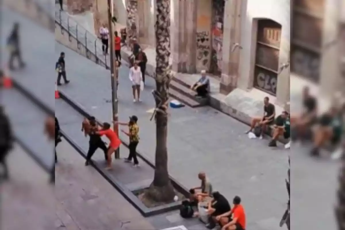 Una escena urbana mostra diverses persones interactuant en un carrer, amb alguns asseguts a terra i altres drets, mentre dos individus semblen estar en una confrontació física a prop d'una palmera.