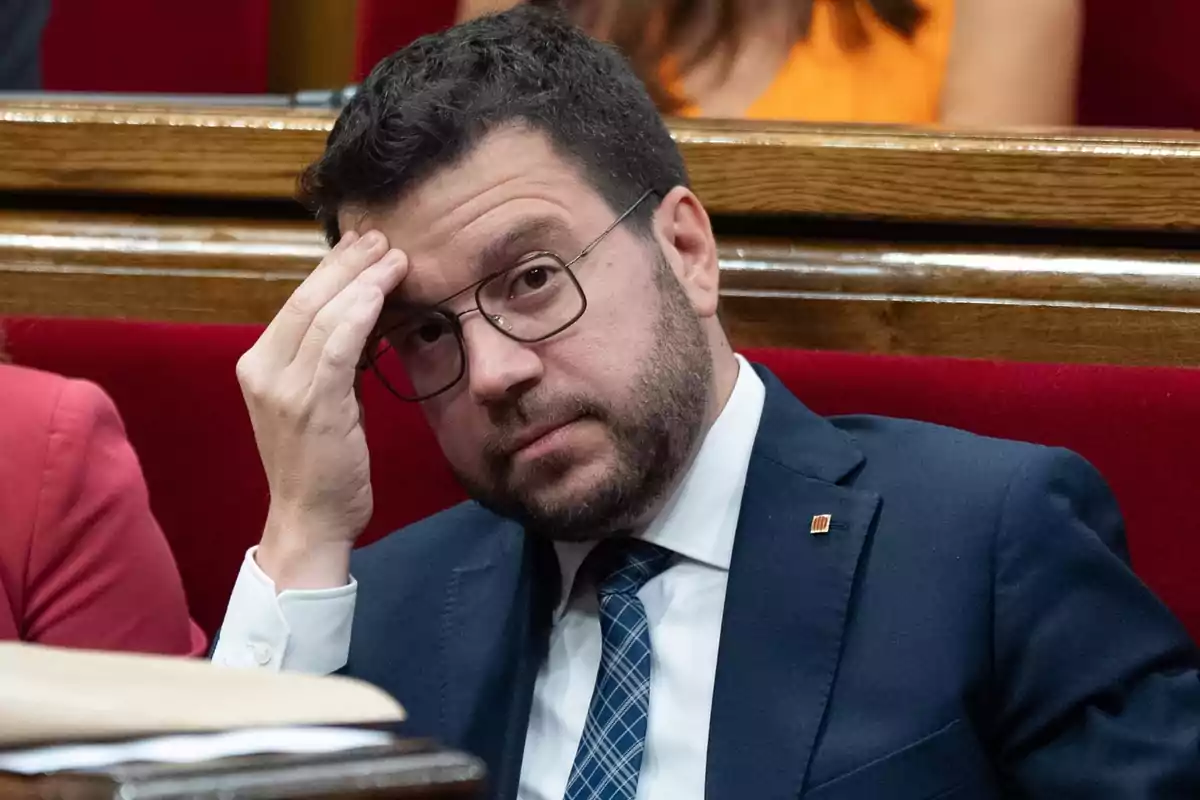 Un hombre con gafas y traje oscuro se toca la frente con una expresión pensativa mientras está sentado en un asiento rojo.