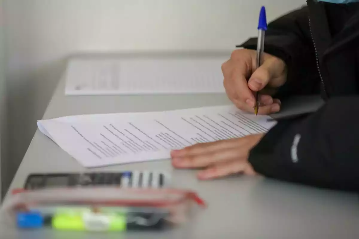 Una persona escrivint en un document sobre una taula amb un bolígraf blau, al costat d'una calculadora i un estoig. Una persona escrivint en un document sobre una taula amb un bolígraf blau, al costat d'una calculadora i un estoig.