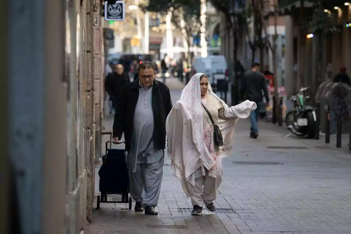 Una parella de gent gran caminant per un carrer urbà, l'home porta una maleta amb rodes i la dona vesteix una vestimenta tradicional.