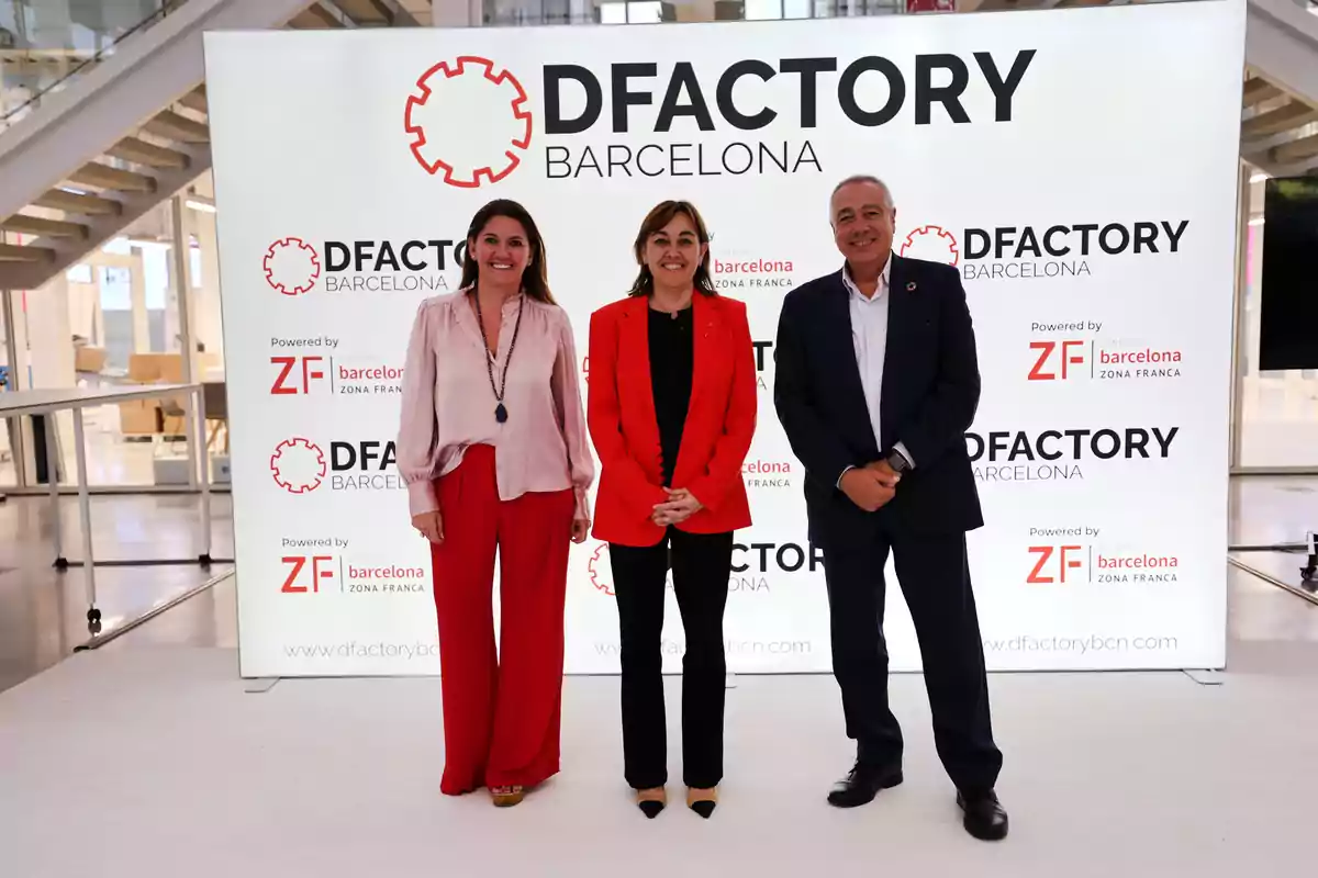 Tres persones posen davant d'un cartell de DFactory Barcelona en un esdeveniment.