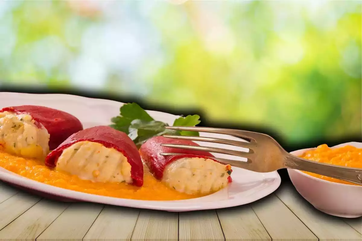 Muntatge plat amb pebrots farcits de bacallà amb una forquilla a sobre al costat d'un bol amb més salsa sobre fons d'imatge d'una taula de fusta en un jardí desenfocada