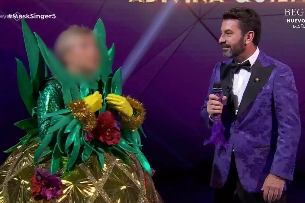 Una persona disfressada de pinya amb fulles verdes i flors és al costat d'Arturo Valls que porta un vestit morat en un escenari de Mask Singer.