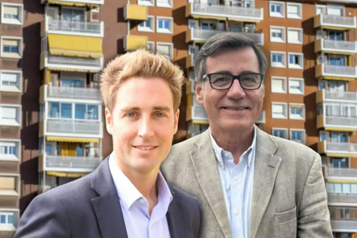 Dos hombres posan frente a un edificio de apartamentos con balcones y toldos amarillos.