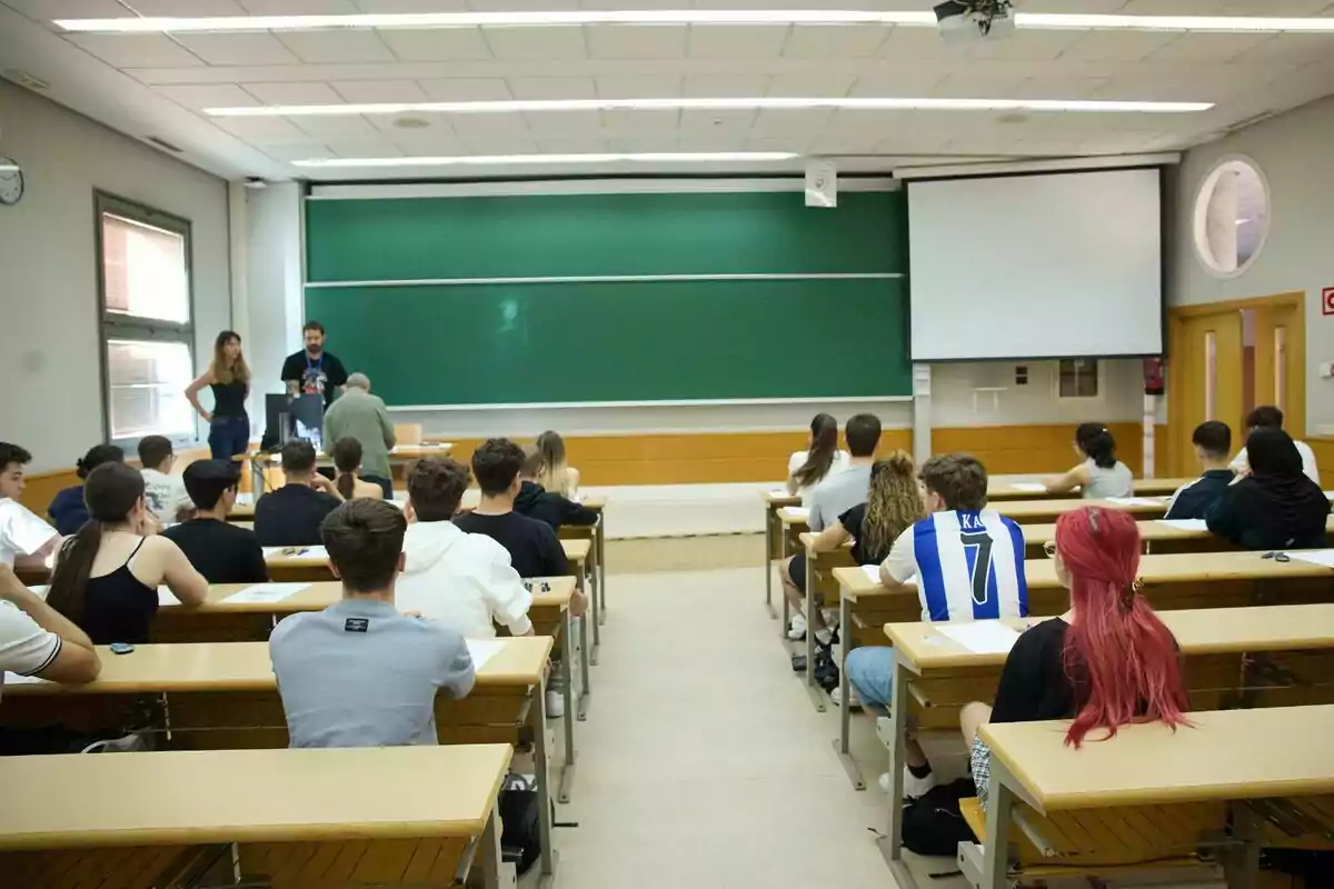 Aula universitària amb estudiants asseguts als pupitres mirant cap al davant, on tres persones estan dretes al costat d’una pissarra verda i una pantalla de projecció. Aula universitària amb estudiants asseguts als pupitres mirant cap al davant, on tres persones estan dretes al costat d’una pissarra verda i una pantalla de projecció.