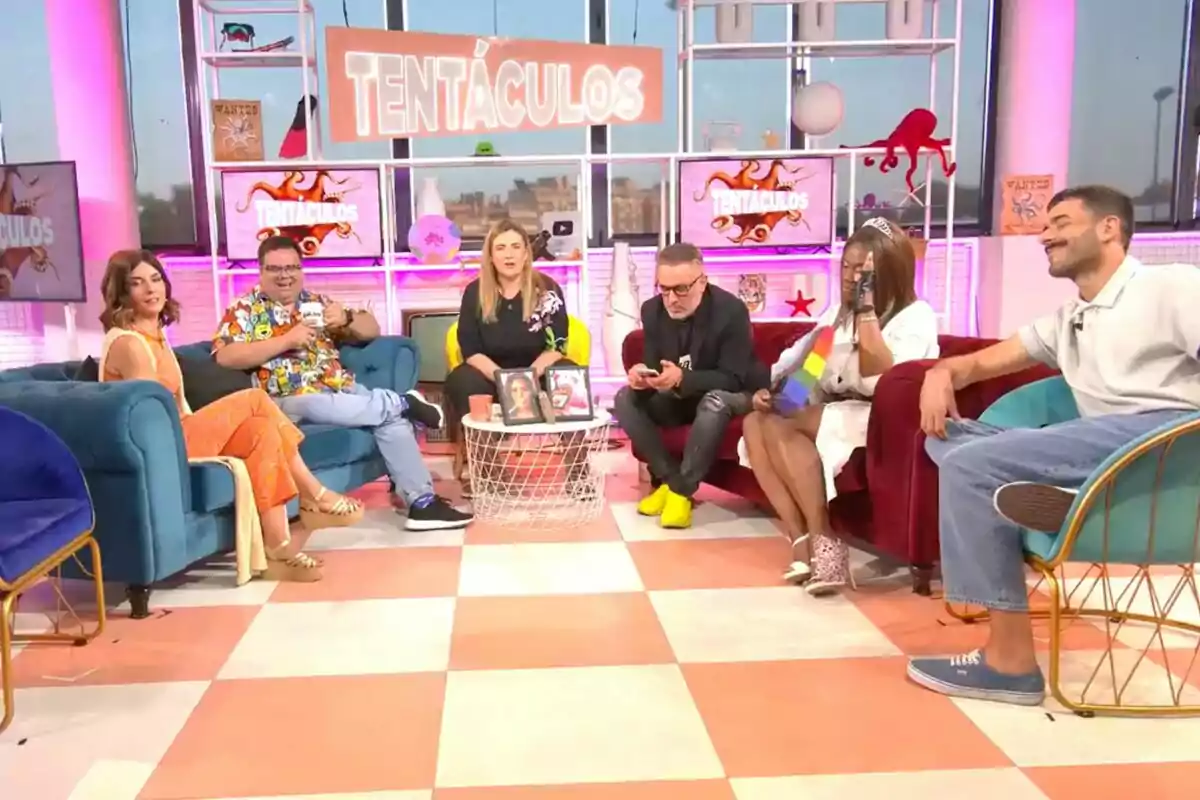 Sis persones estan assegudes en un plató de televisió acolorit, 'Tentacles', decorat amb temàtica de tentacles, conversant i somrient en un ambient relaxat. Sis persones estan assegudes en un plató de televisió acolorit, 'Tentacles', decorat amb temàtica de tentacles, conversant i somrient en un ambient relaxat.
