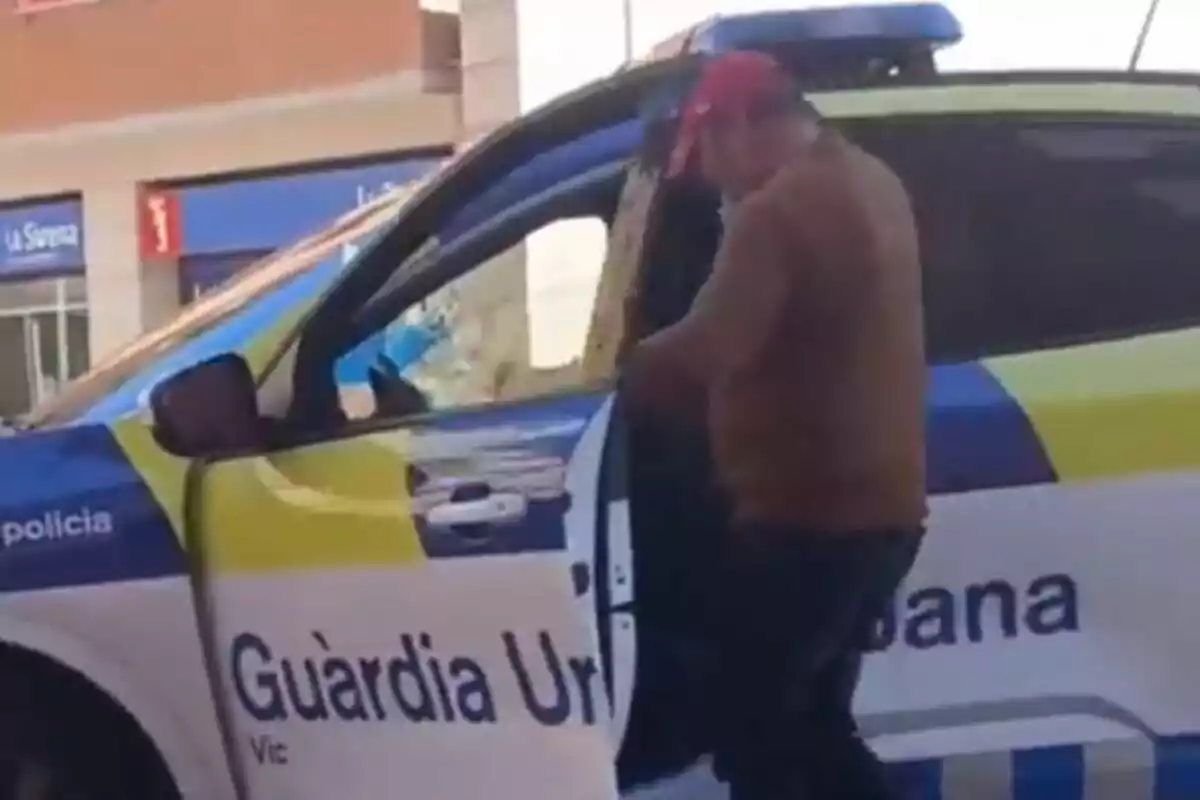 Un home amb gorra vermella és dret al costat d'un cotxe de la Guàrdia Urbana de Vic, amb la porta del vehicle oberta.