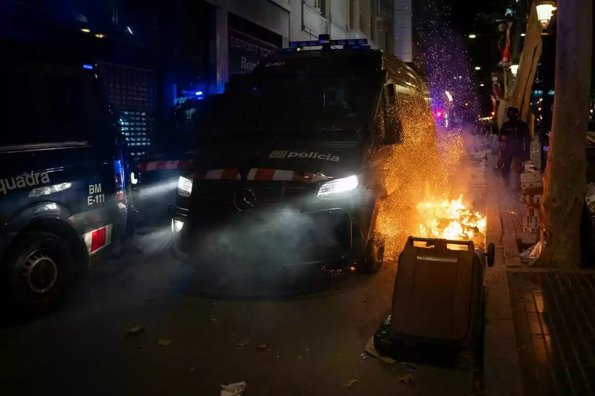 Una furgoneta de policía junto a un contenedor en llamas en una calle por la noche.