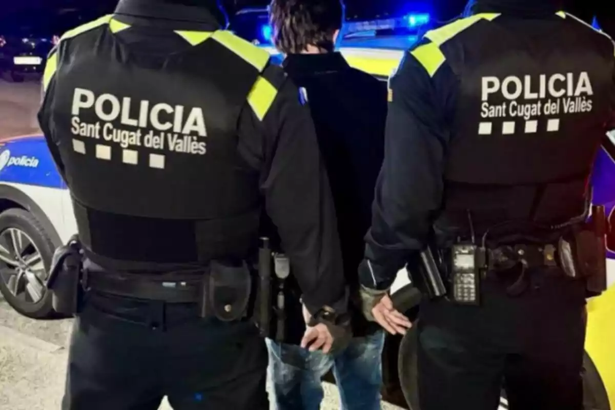 Dos policies de Sant Cugat del Vallès escorten una persona emmanillada davant d'un cotxe patrulla.