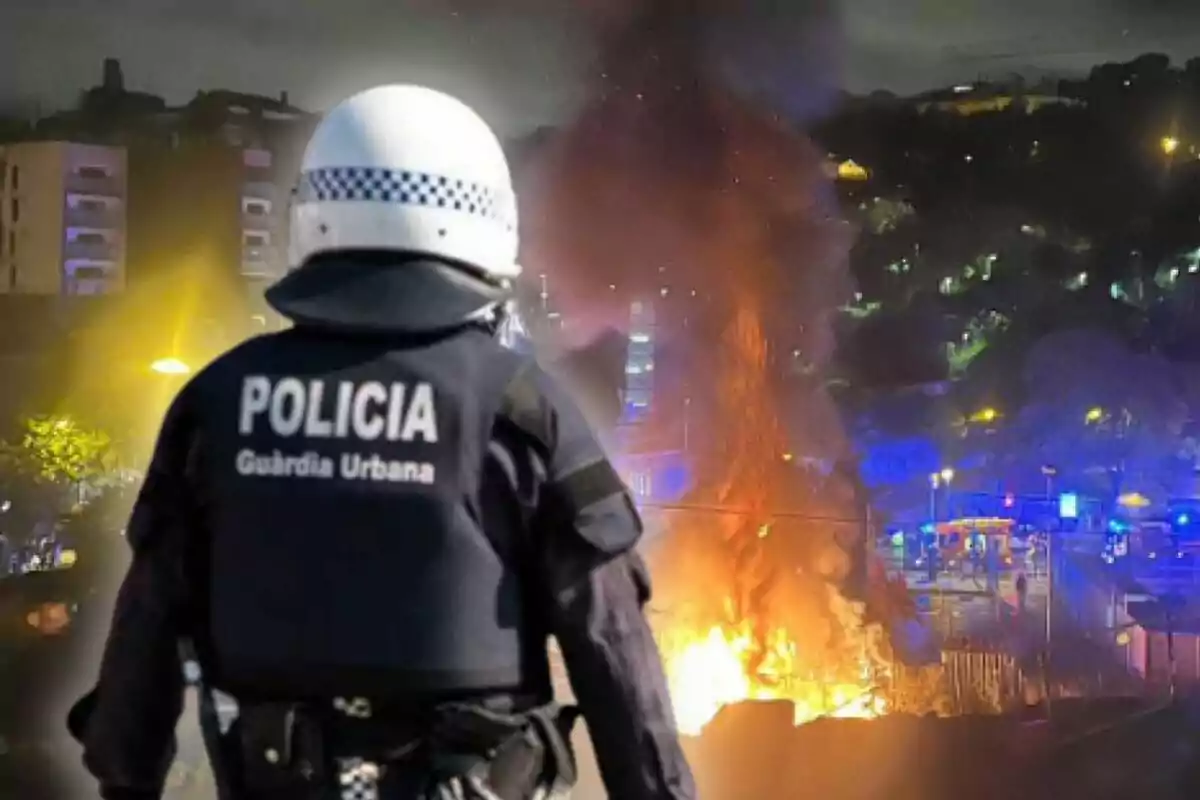 Un agent de la policia de la Guàrdia Urbana observa un incendi a la ciutat durant la nit, amb flames i fum elevant-se al fons.