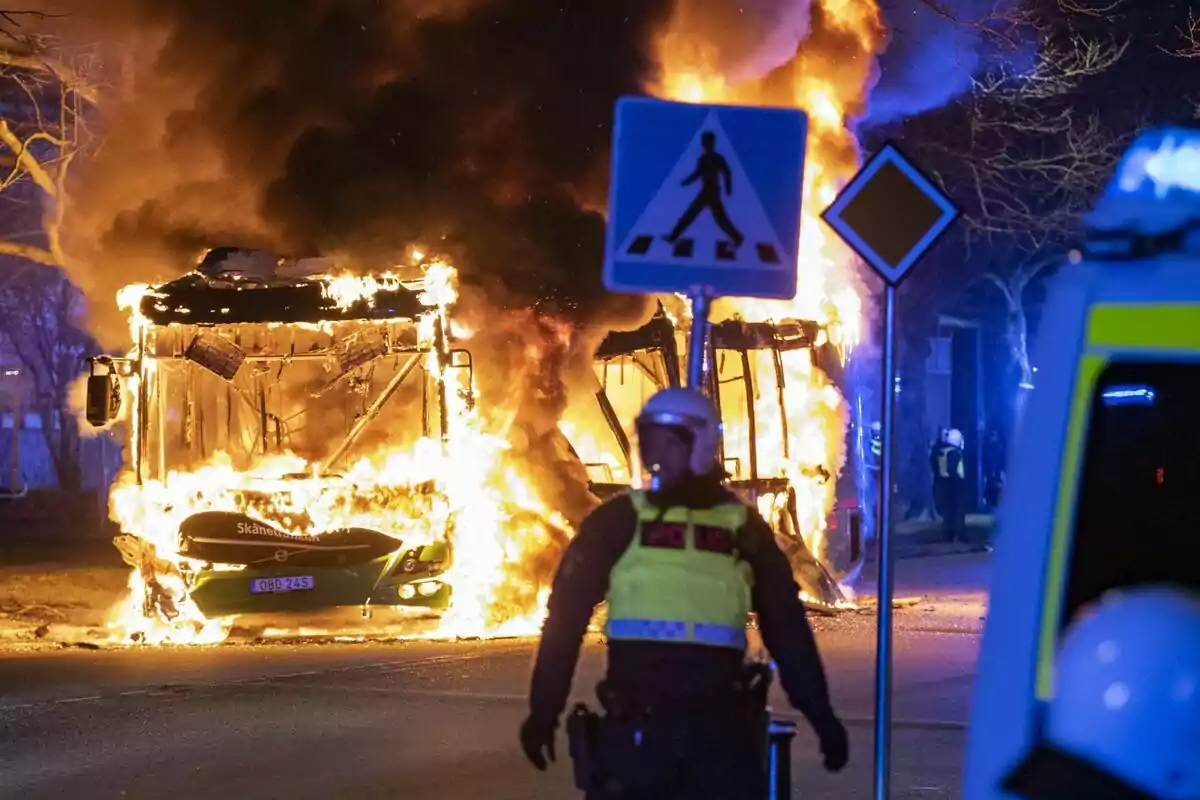 Un autobús en flames en un carrer durant la nit amb un policia en primer pla i senyals de trànsit visibles. Un autobús en flames en un carrer durant la nit amb un policia en primer pla i senyals de trànsit visibles.