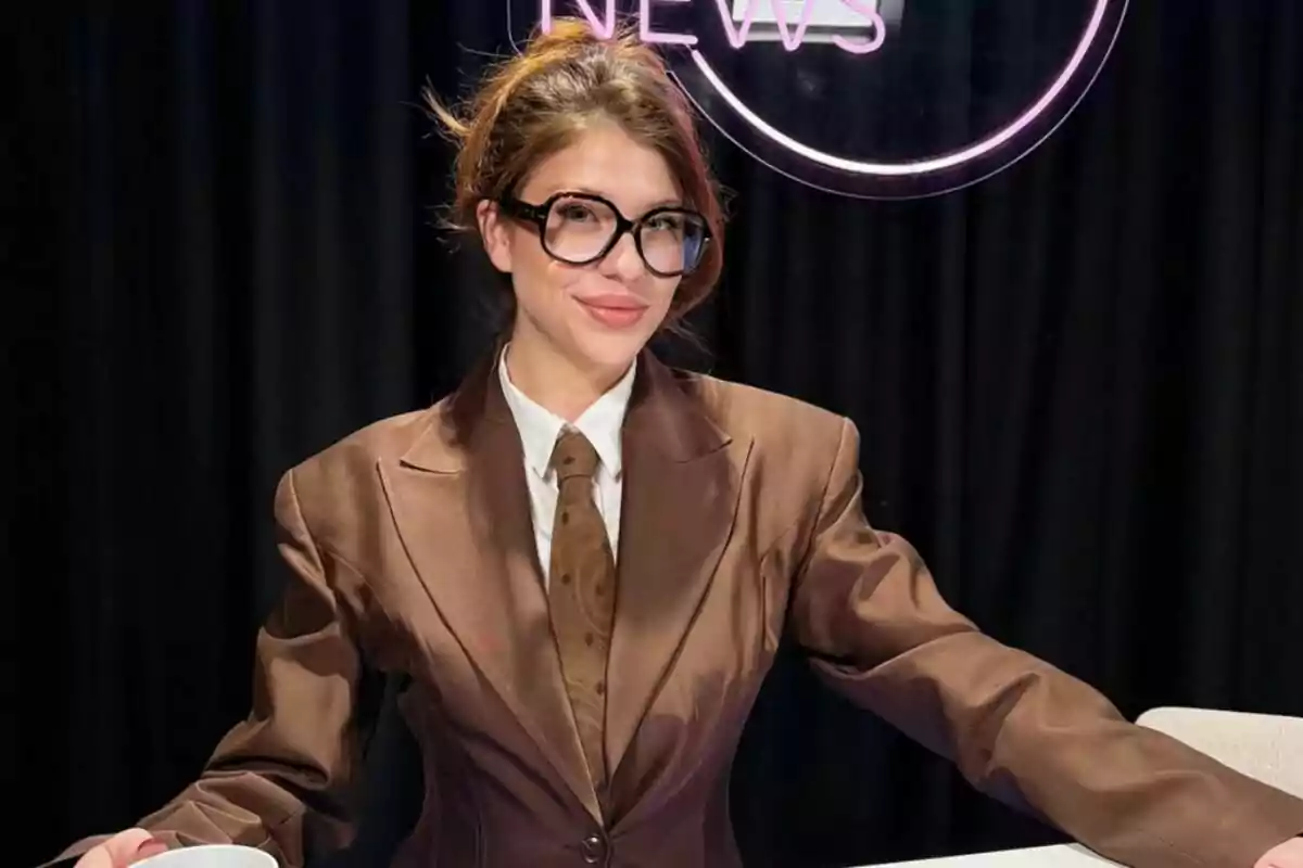 Anita Matamoros con gafas y traje marrón está sentada frente a un fondo oscuro con un letrero de neón que dice "Show Business NEWS".