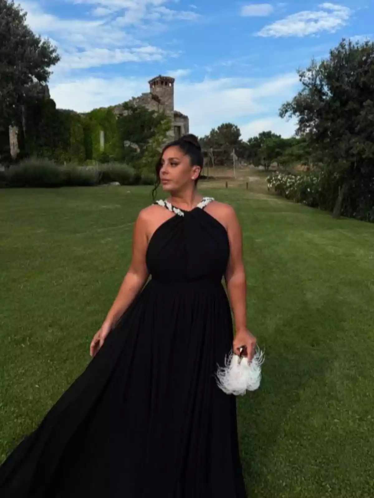 Anabel Pantoja in a long black dress and a white feathered bag posing in a garden with ruins covered in vegetation in the background under a blue sky with clouds Anabel Pantoja in a long black dress and a white feathered bag posing in a garden with ruins covered in vegetation in the background under a blue sky with clouds