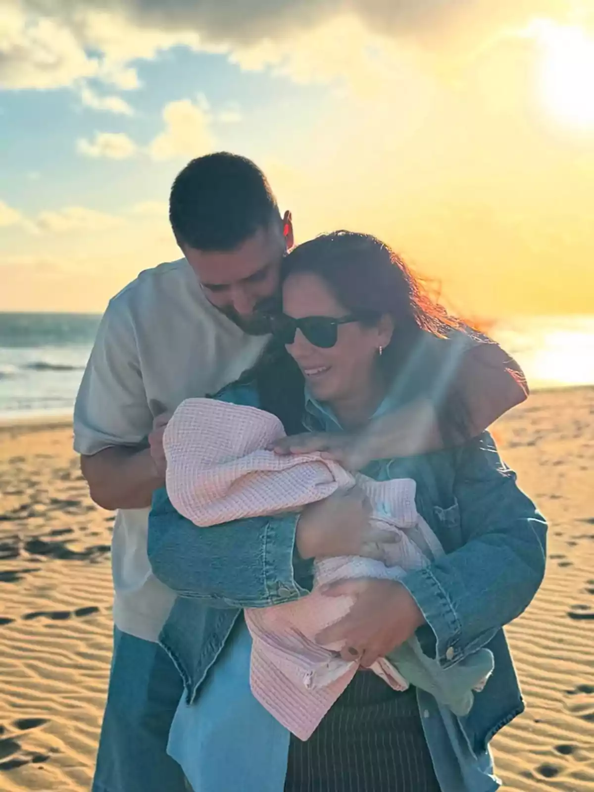 David Rodríguez and a smiling Anabel Pantoja hold Alma wrapped in a blanket on the beach at sunset.