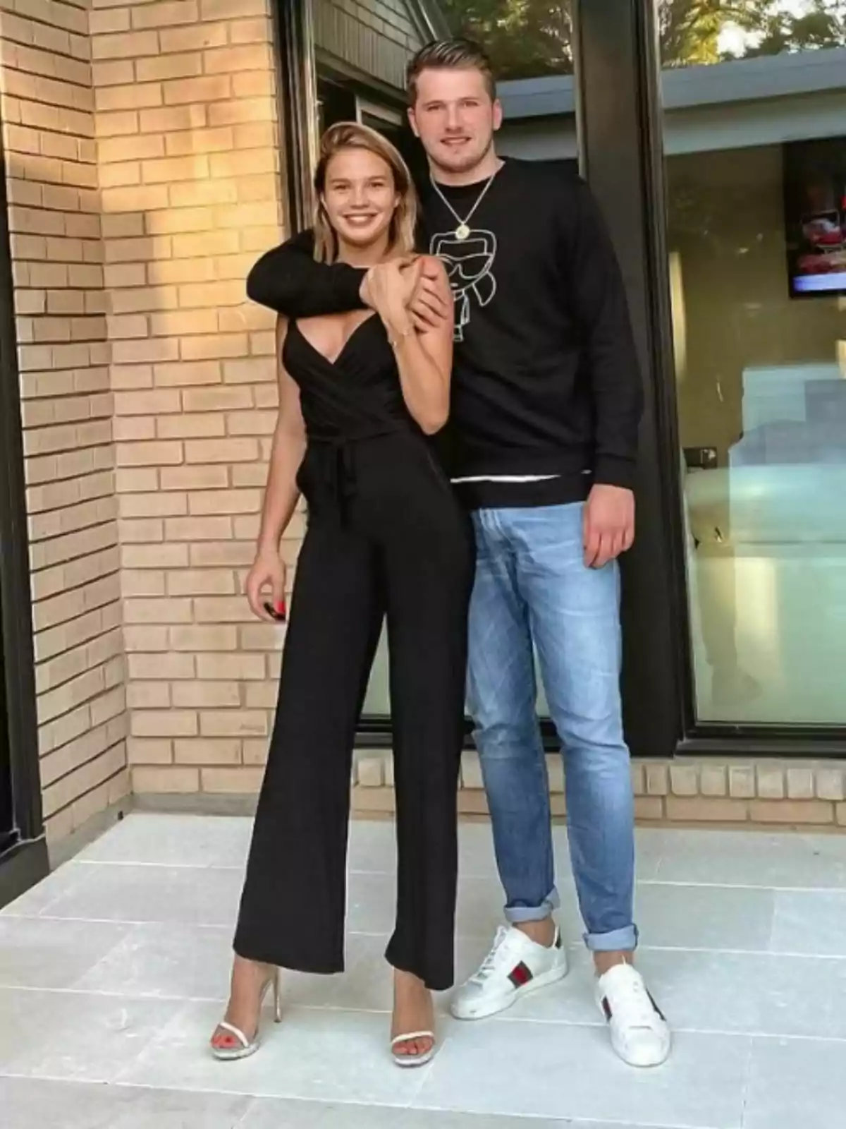 Anamaria Goltes and Luka Doncic posing together in front of a brick wall, she is wearing a black jumpsuit and heels, he is wearing a black sweater and jeans.