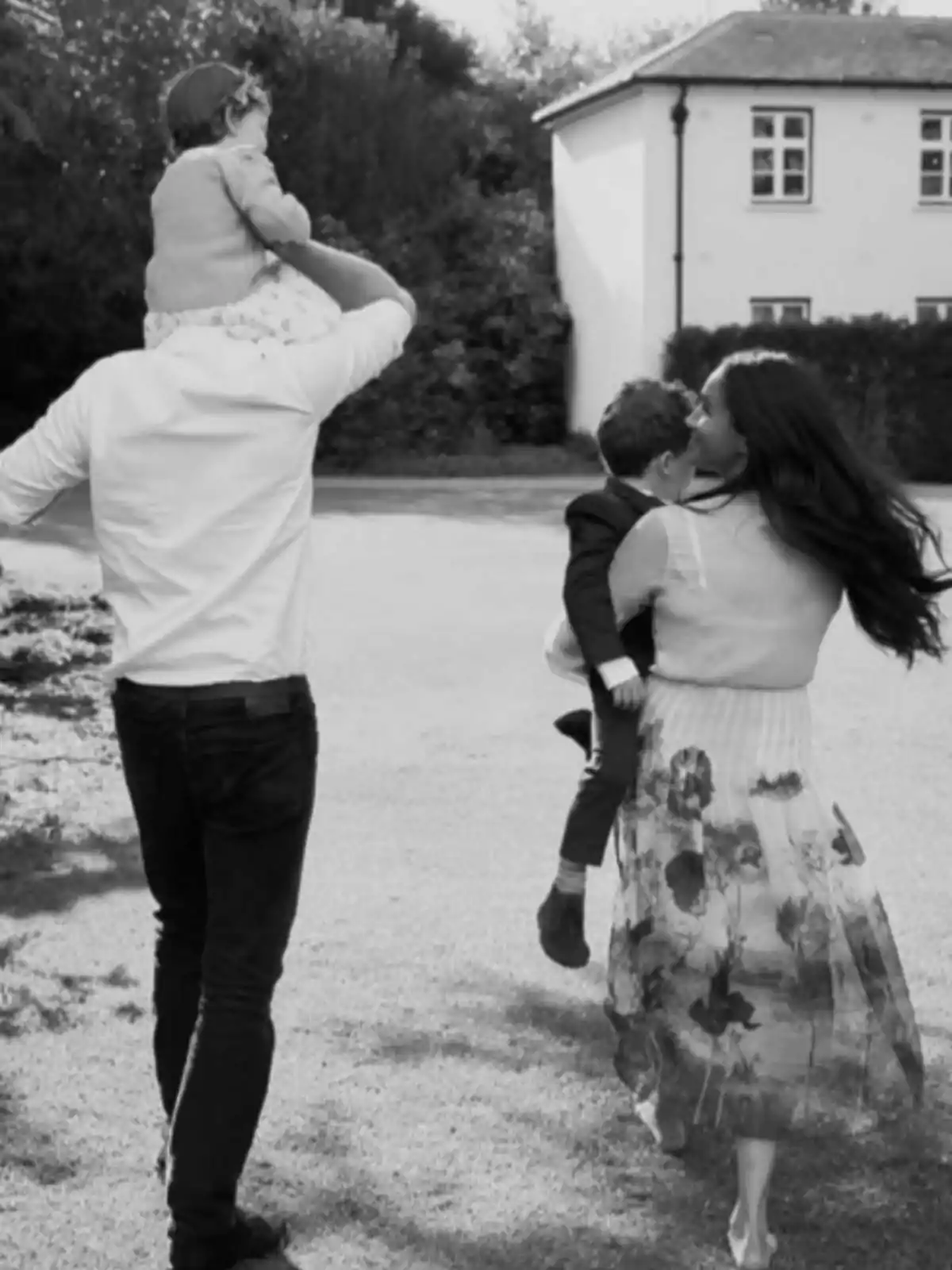 A family walking outdoors, with a man carrying a girl on his shoulders and a woman holding a boy in her arms, as they head toward a house.