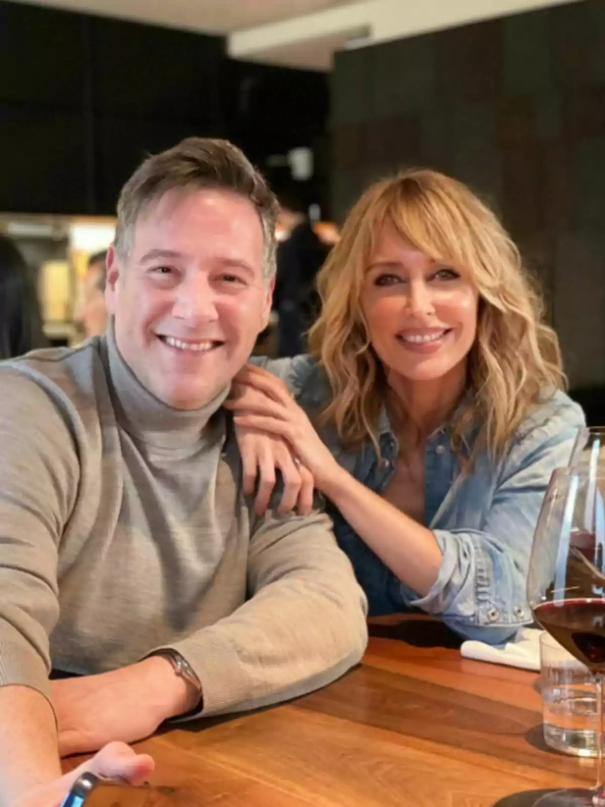 Carlos Latre and Emma García smile while sitting at a wooden table in a restaurant with wine glasses. Carlos Latre and Emma García smile while sitting at a wooden table in a restaurant with wine glasses.