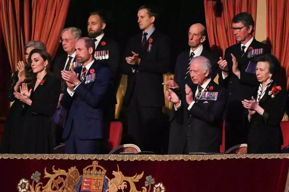 La família reial britànica vestides formalment aplaudeix en un esdeveniment, amb cortines vermelles de fons i un escut decoratiu en primer pla. La família reial britànica vestides formalment aplaudeix en un esdeveniment, amb cortines vermelles de fons i un escut decoratiu en primer pla.