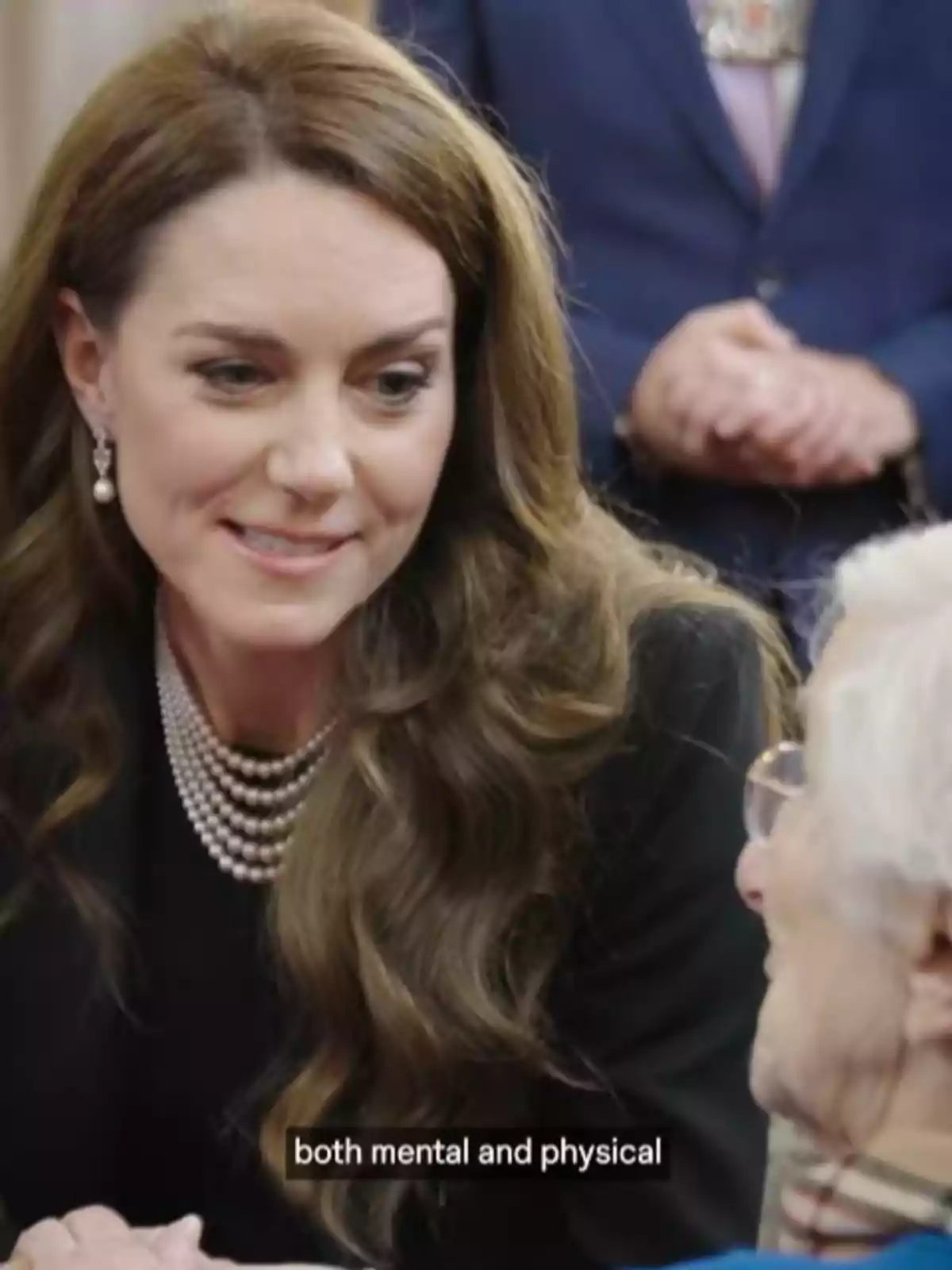 Kate Middleton with brown hair and pearls smiles while talking to an older person wearing glasses.