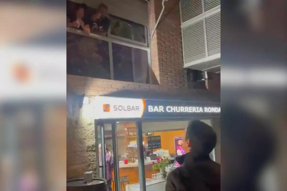 Una persona mira cap a una finestra des del carrer mentre altres persones s'aboquen des de l'interior d'un edifici sobre un bar xurreria anomenat Solbar.