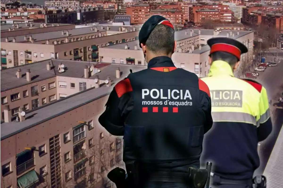 Two police officers with their backs turned, wearing Mossos d'Esquadra uniforms, in front of a group of residential buildings.