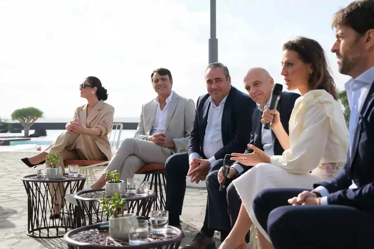 Un grup de persones assegudes a l'aire lliure participant en una conversa amb taules petites i plantes en primer pla.