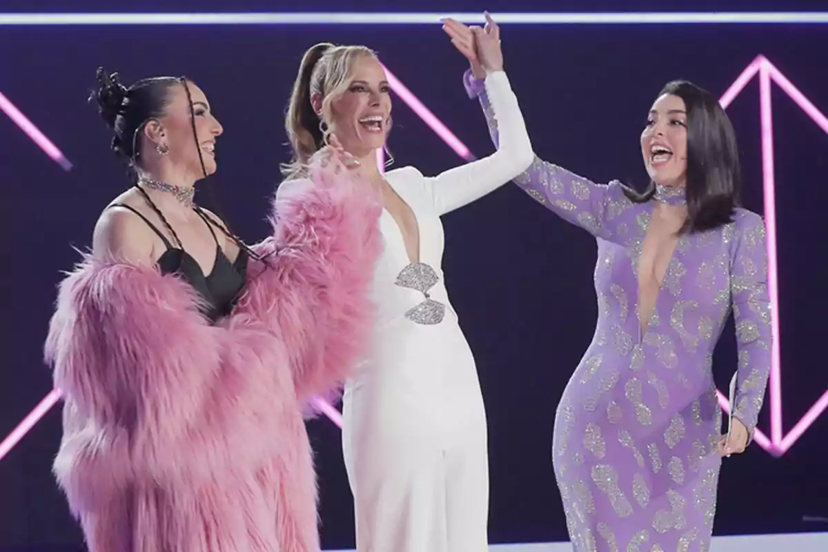 Inés Hernand, Paula Vázquez i Ruth Lorenzo posen juntes en un escenari del Benidorm Fest amb llums de neó, una porta un abric rosa pelut, una altra un vestit blanc i la tercera un vestit morat amb detalls brillants.