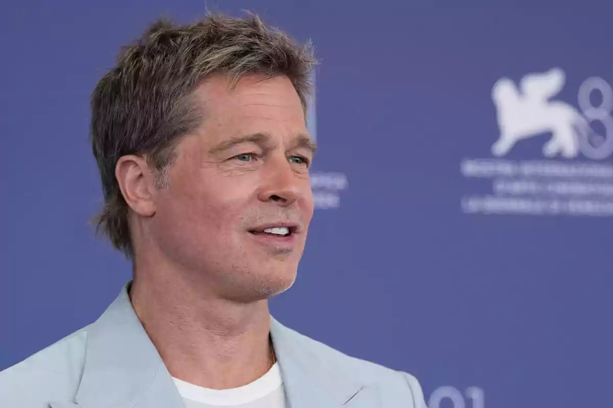 Brad Pitt in a light blue jacket is smiling in front of a blue background with a white logo. Brad Pitt in a light blue jacket is smiling in front of a blue background with a white logo.