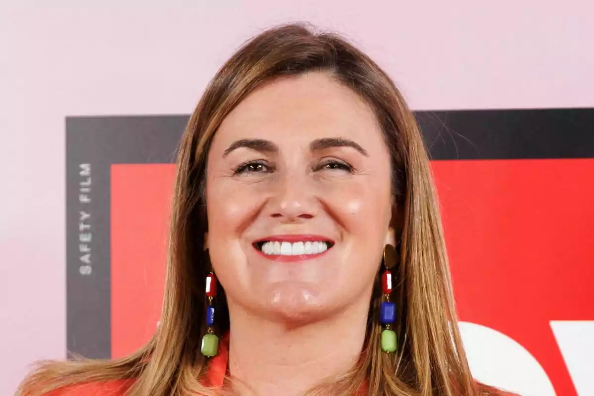 Carlota Corredera sonriendo con pendientes coloridos frente a un fondo rojo y negro.