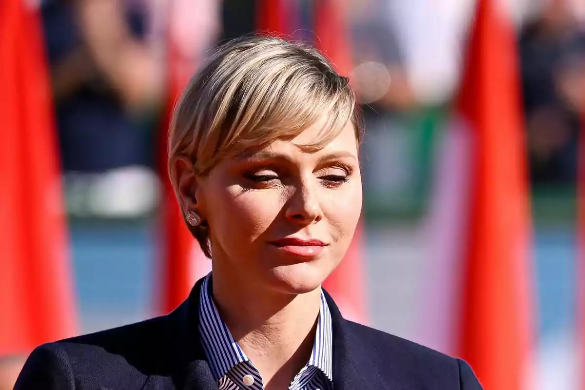 Charlene con cabello corto y rubio, ojos cerrados y expresión serena, vestida con una camisa a rayas y un saco oscuro, con un fondo de banderas rojas desenfocadas. Charlene con cabello corto y rubio, ojos cerrados y expresión serena, vestida con una camisa a rayas y un saco oscuro, con un fondo de banderas rojas desenfocadas.