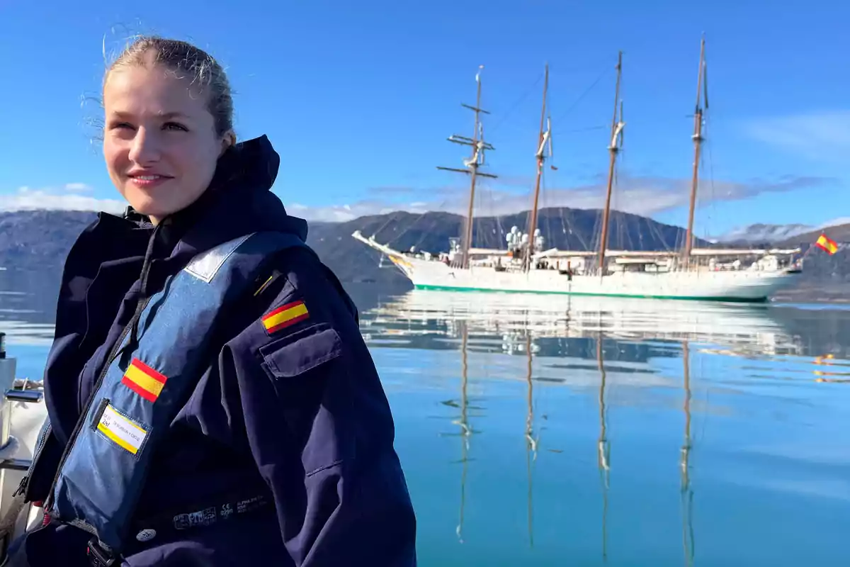 Princess Leonor in a navy blue uniform and Spanish flags on the life jacket is on a boat with a sailboat in the background in a mountainous landscape and clear sky. Princess Leonor in a navy blue uniform and Spanish flags on the life jacket is on a boat with a sailboat in the background in a mountainous landscape and clear sky.