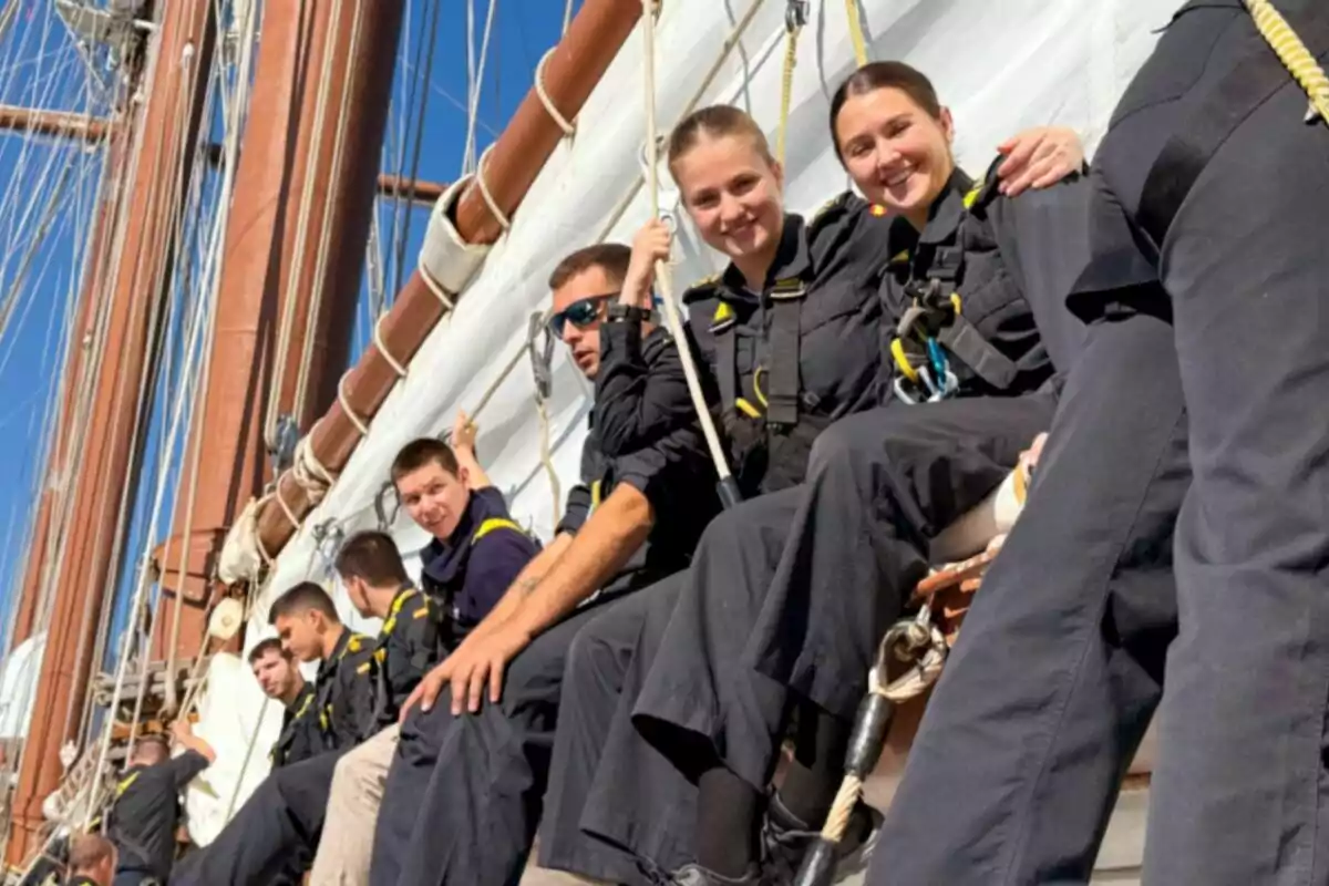 A group of people dressed in dark uniforms is sitting on the structure of a sailing ship, holding onto ropes and smiling as they enjoy the sunny day. A group of people dressed in dark uniforms is sitting on the structure of a sailing ship, holding onto ropes and smiling as they enjoy the sunny day.