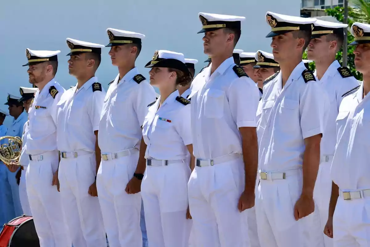 Un grupo de personas vestidas con uniformes blancos de la marina están formados en fila durante una ceremonia al aire libre.