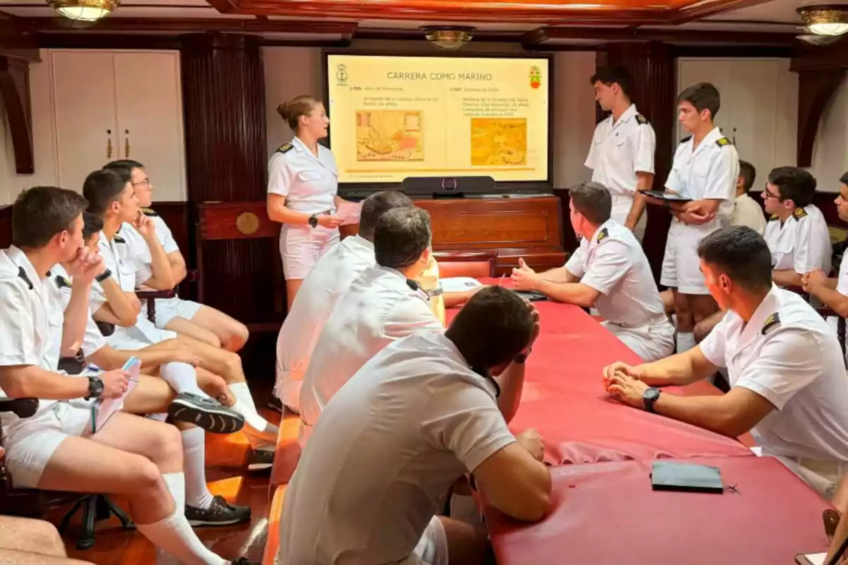 A group of people in naval uniforms attends a presentation in a room with a screen displaying information about a career as a sailor. A group of people in naval uniforms attends a presentation in a room with a screen displaying information about a career as a sailor.