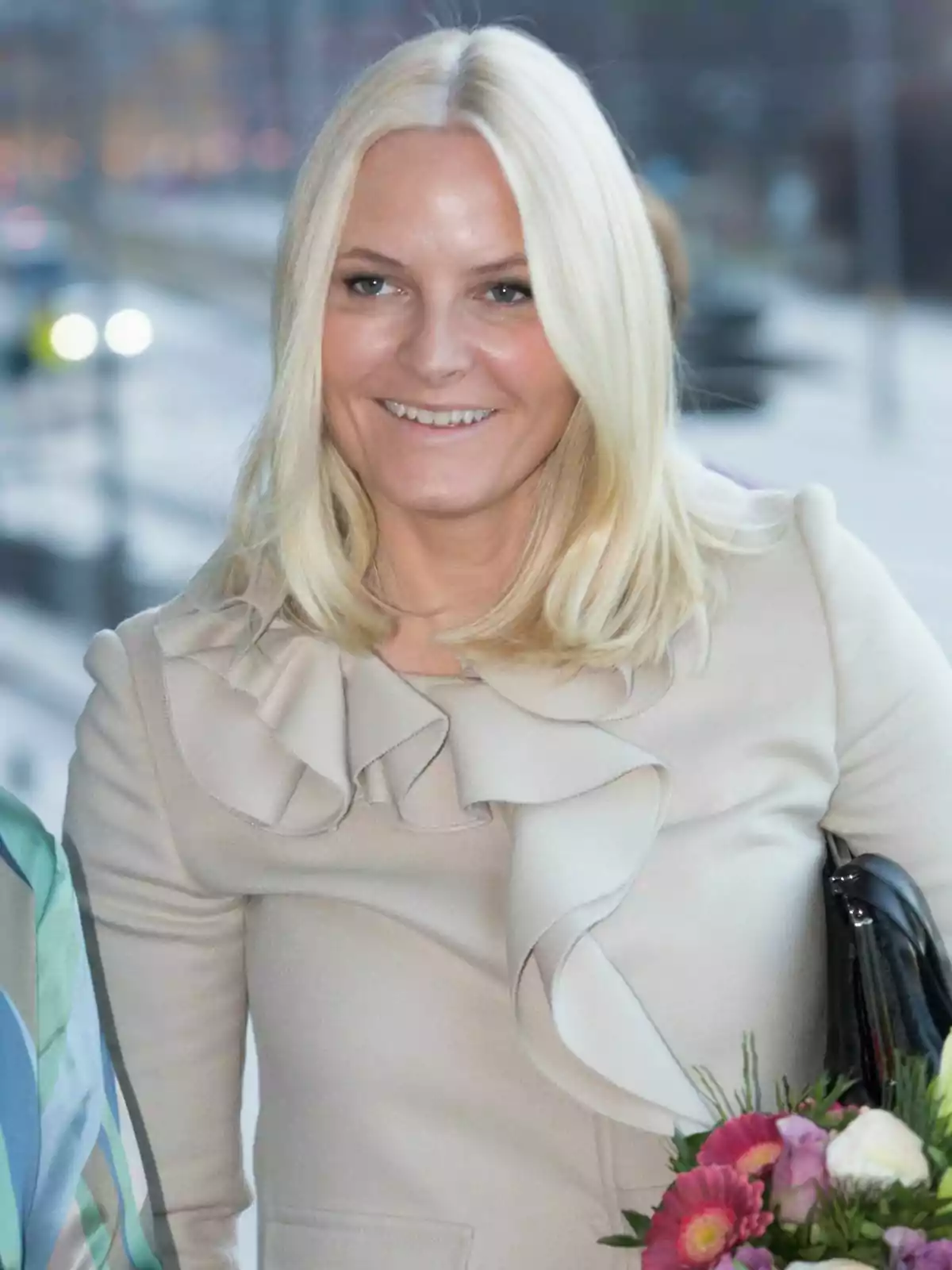 Princess Mette-Marit, wearing a light-coloured coat with ruffled details, holds a bouquet of flowers as she smiles. Princess Mette-Marit, wearing a light-coloured coat with ruffled details, holds a bouquet of flowers as she smiles.