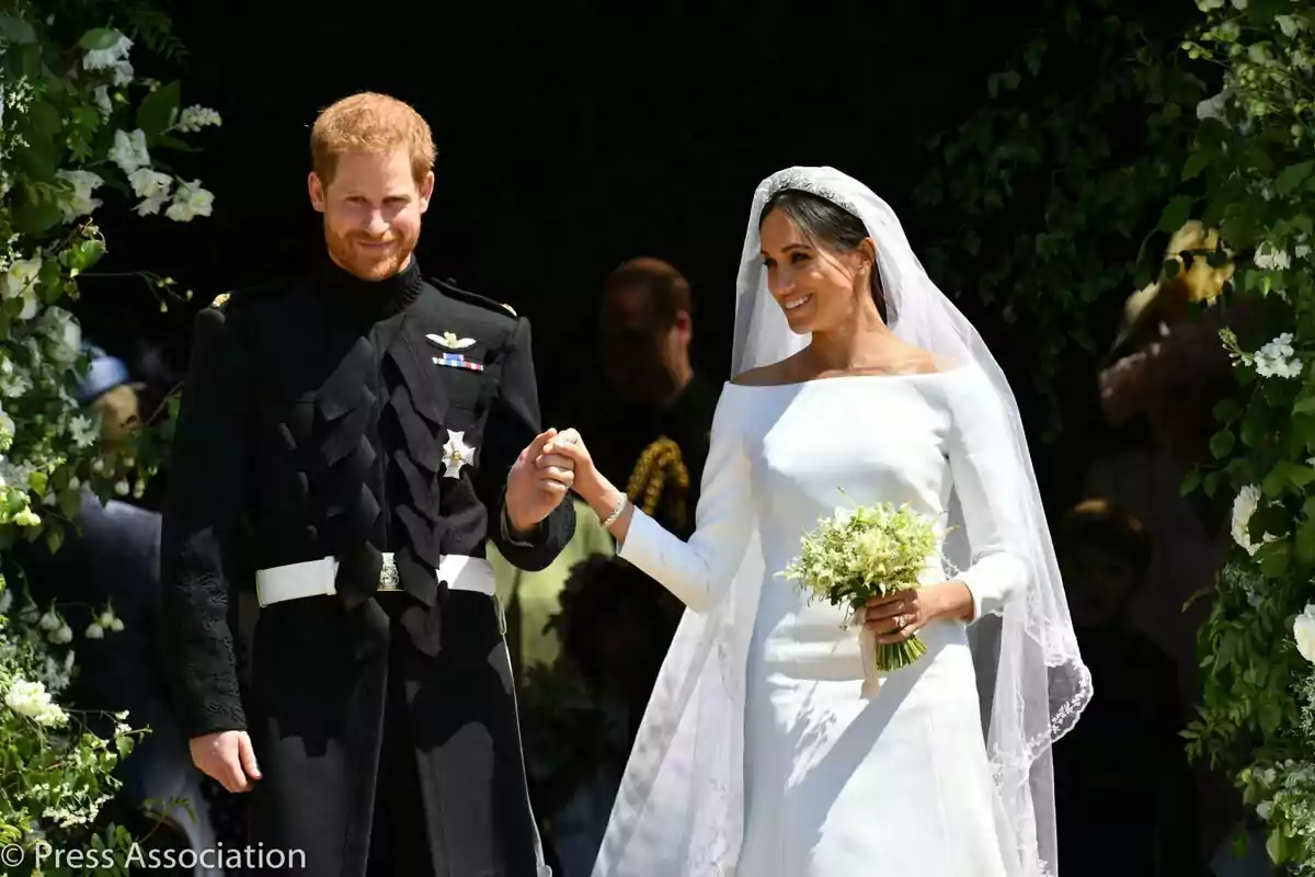 Prince Harry and Meghan Markle smiling in wedding dress leave a building decorated with flowers.