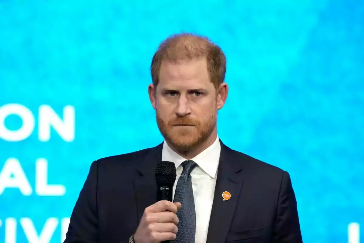 Prince Harry with a beard and dark suit holding a microphone in front of a blue background.