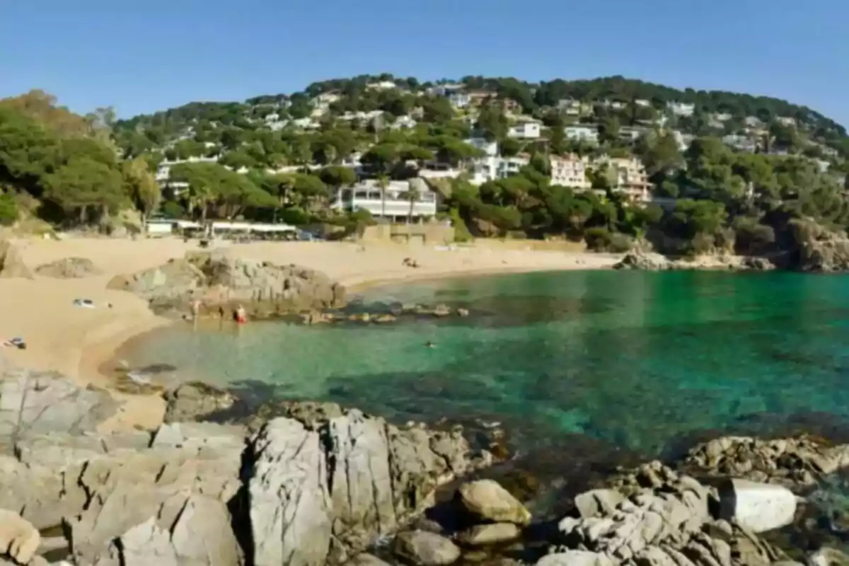 Platja amb aigües cristal·lines envoltada de turons amb cases i vegetació.