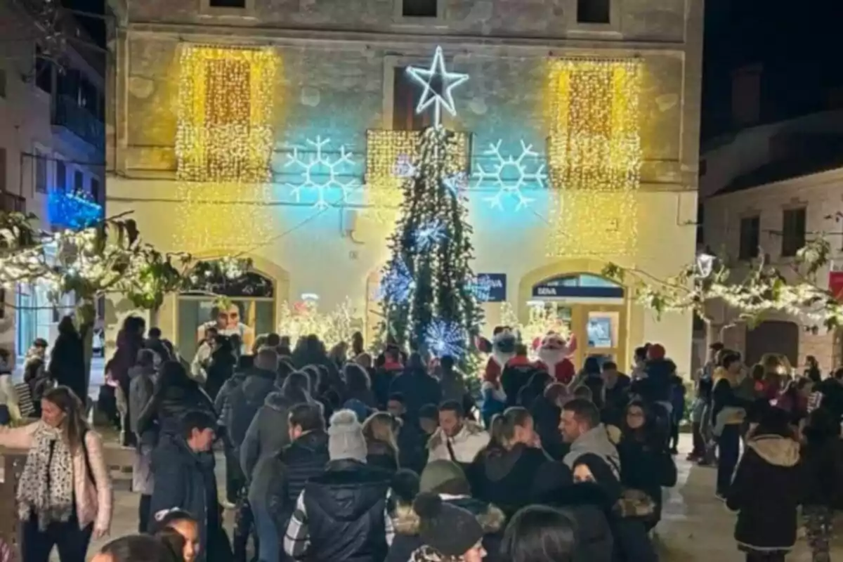 Una multitud de persones es reuneix en una plaça decorada amb llums nadalenques i un arbre il·luminat davant d'un edifici adornat amb estrelles i flocs de neu.