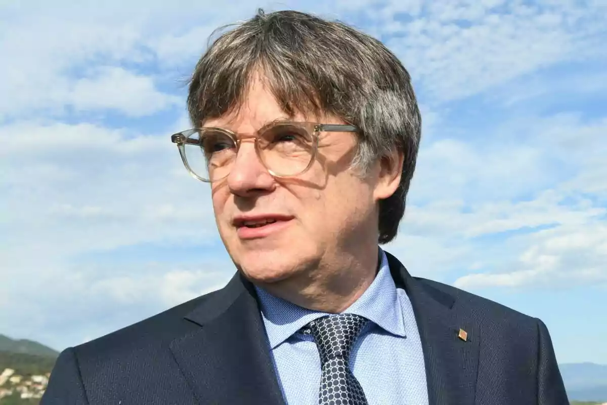 Carles Puigdemont amb ulleres i vestit fosc mirant cap a la dreta amb un cel blau i núvols de fons.