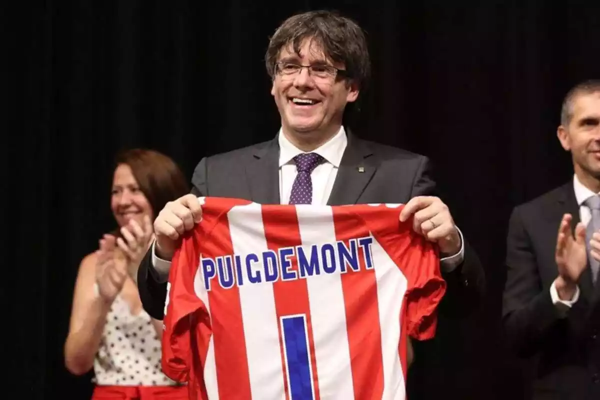 Pla mitjà de Carles Puigdemont somrient i mostrant una samarreta del Girona FC amb el nom de Puigdemont i el dorsal número 1
