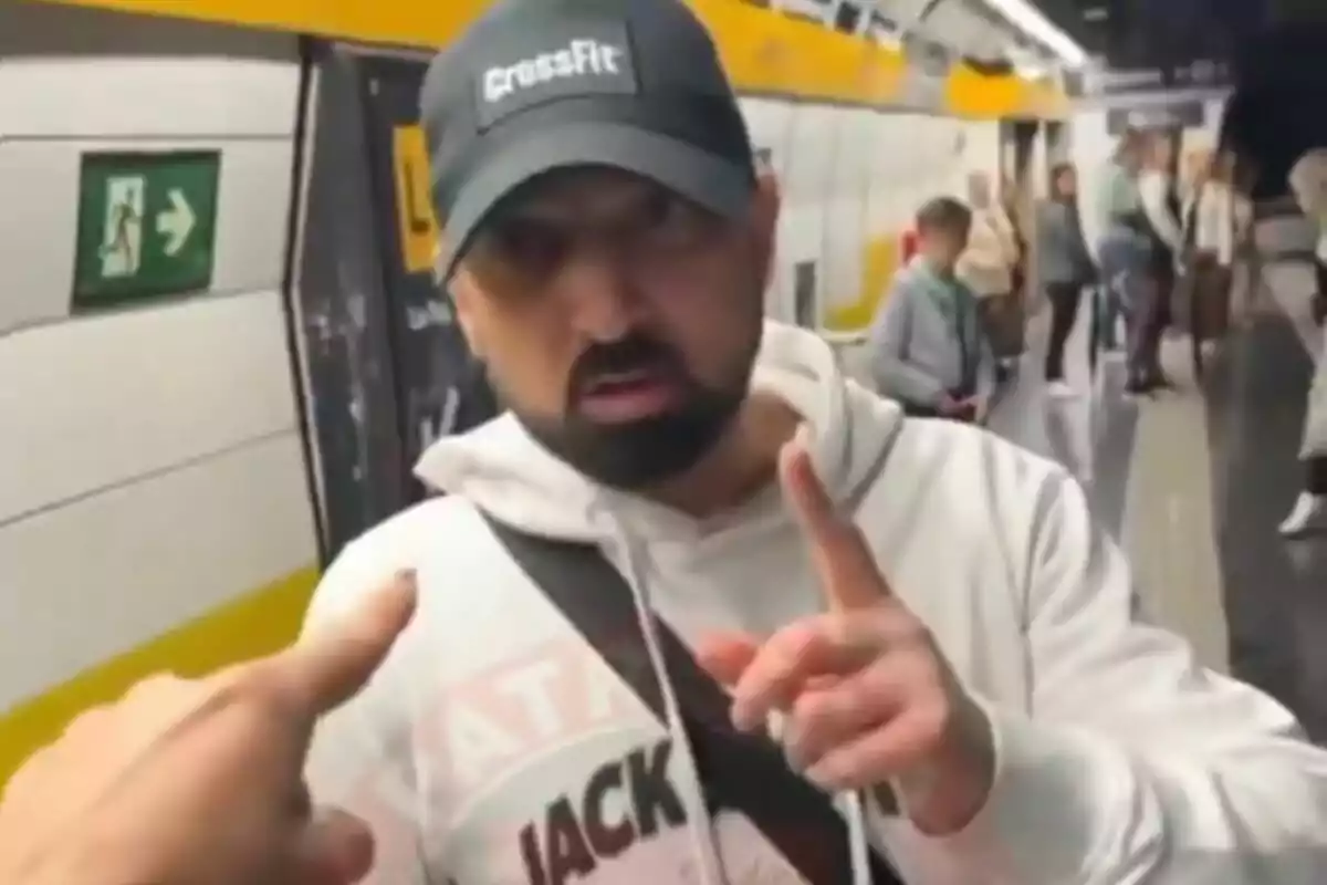 Un home amb gorra i dessuadora blanca està en una estació de metro, assenyalant amb el dit mentre una altra persona ho assenyala a ell.