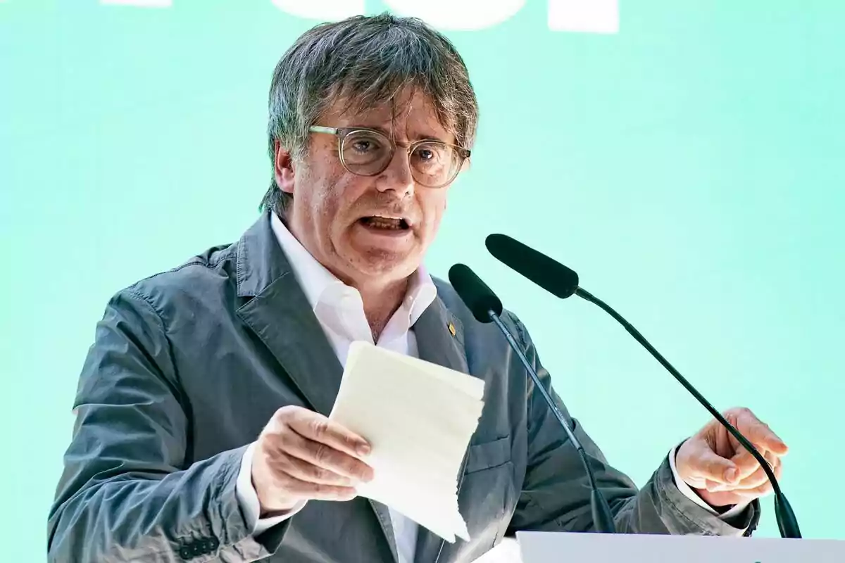 Carles Puigdemont parlant en un podi amb micròfons, sostenint papers a la mà, fons verd.