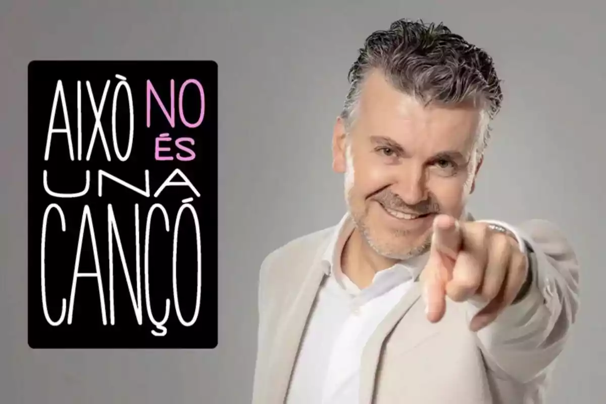 Ramon Gener sonriente con barba y cabello canoso, vestido con un traje claro, señala hacia la cámara; a su lado hay un cartel negro con letras blancas y rosas que dice "Això no és una cançó".