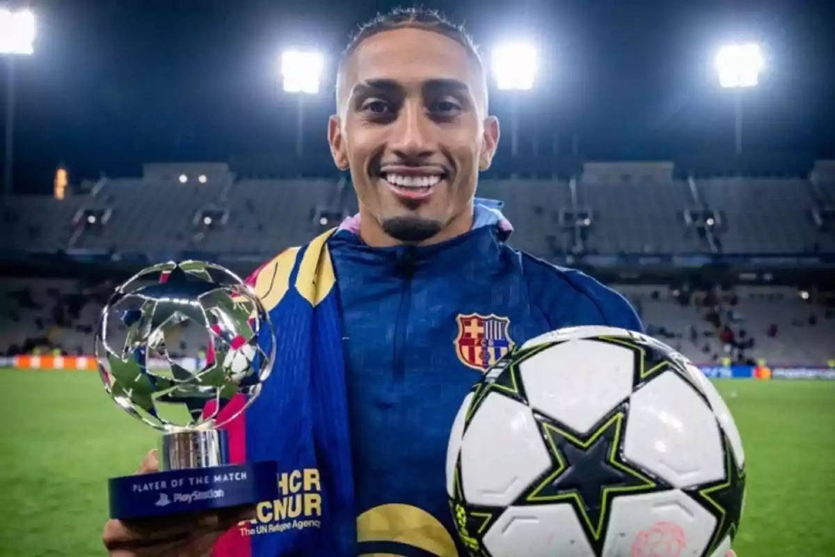 A player smiles while holding a trophy and a ball in a lit-up stadium.