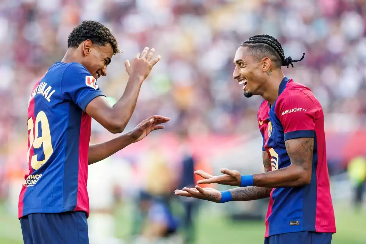 Dos jugadors de futbol amb uniformes blau i vermell somriuen i gesticulen al camp.